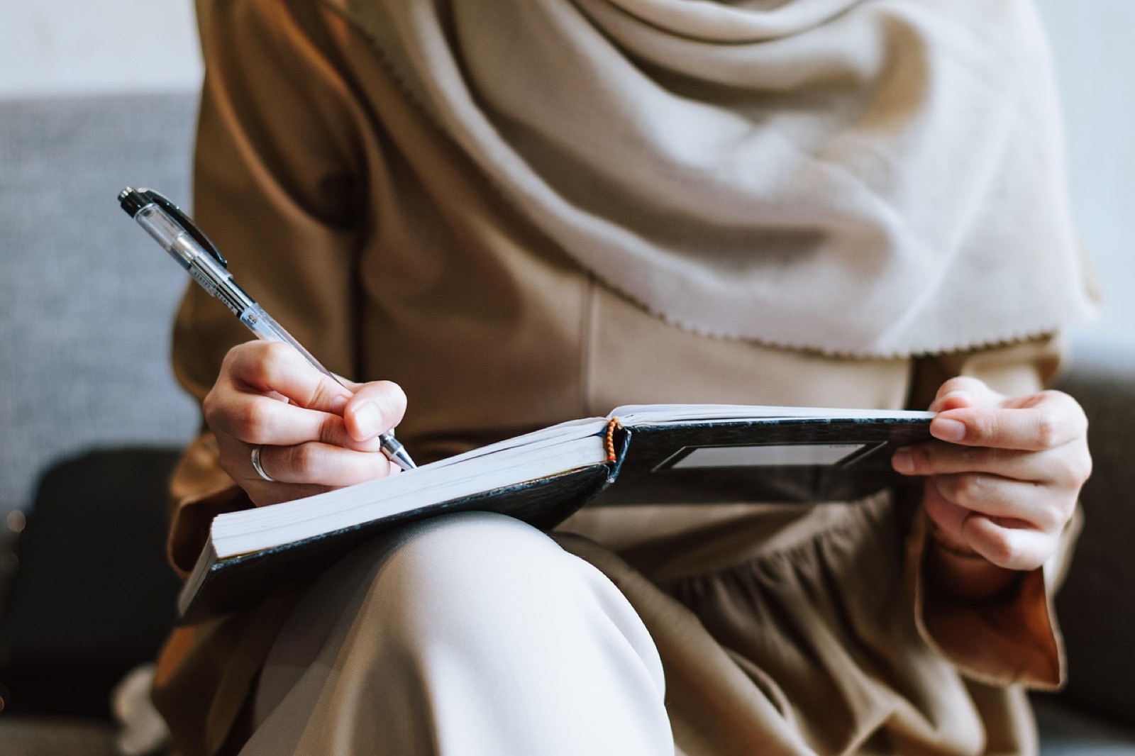 Person in a beige wrap writing in a notebook with a pen