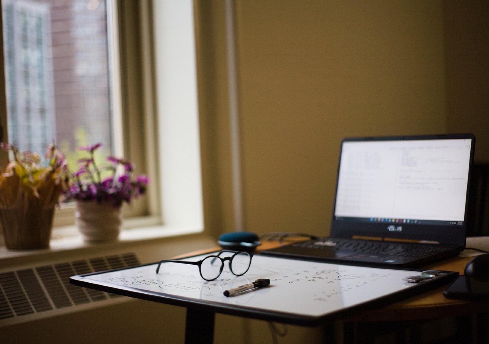 Laptop, eyeglasses and papers on a desk by a sunny window with flowers