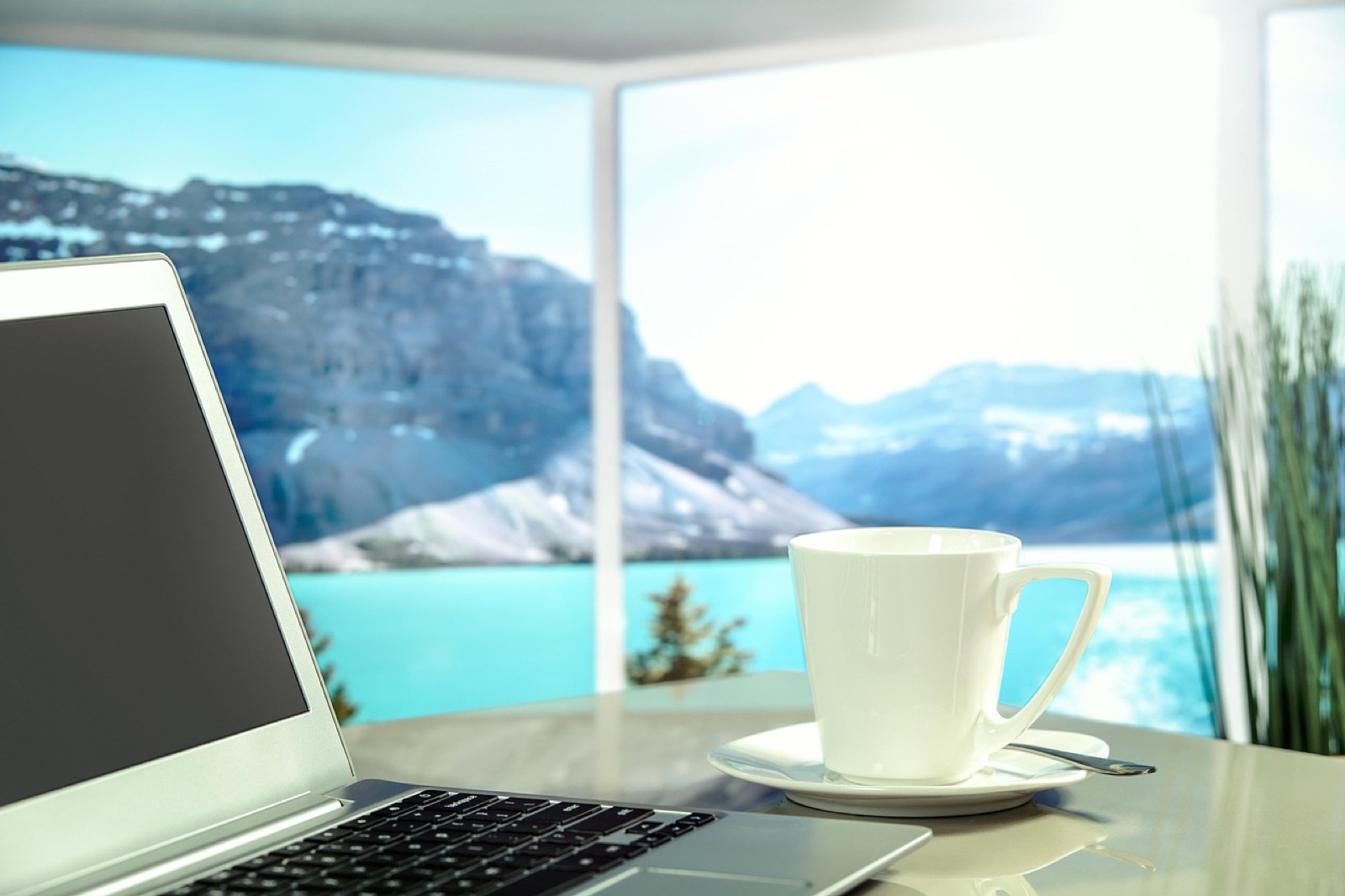 Laptop and white coffee cup on a desk by a window overlooking mountains and a turquoise lake